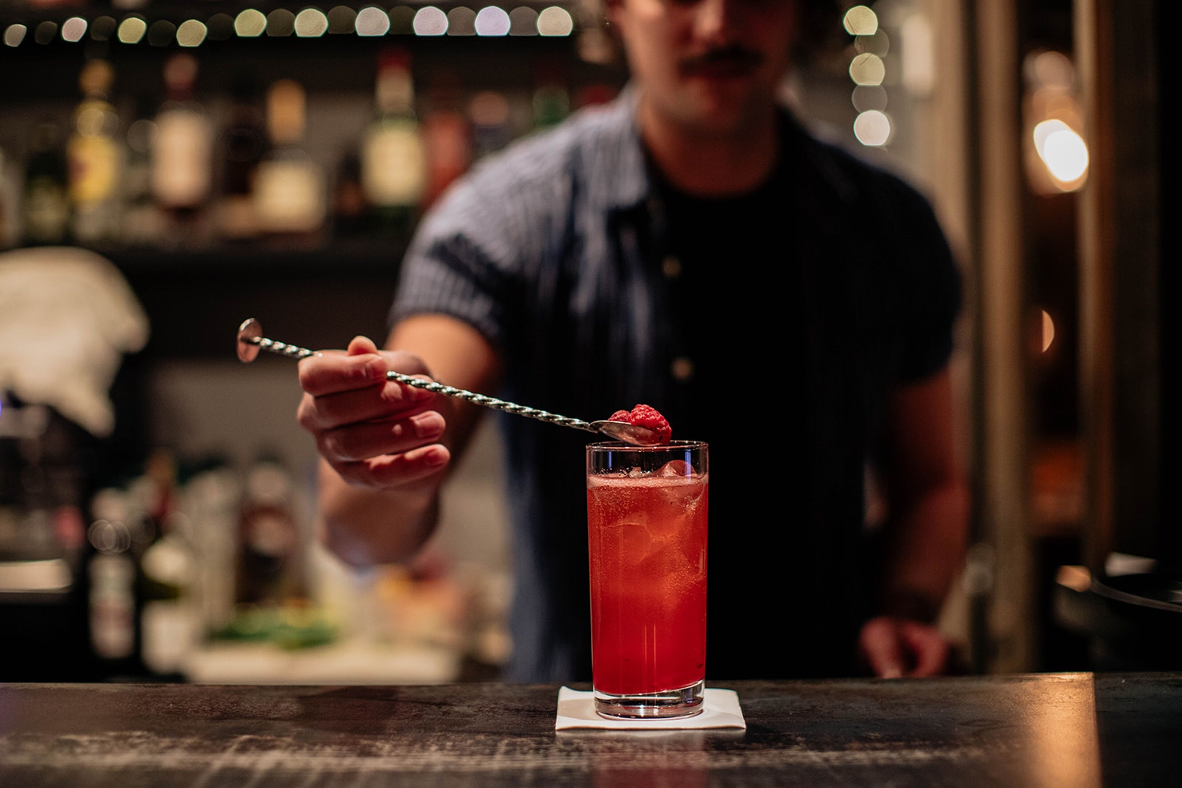 A bar setting with a tall glass of red drink, a spoon with a raspberry, and a blurred background of bottles and a person.