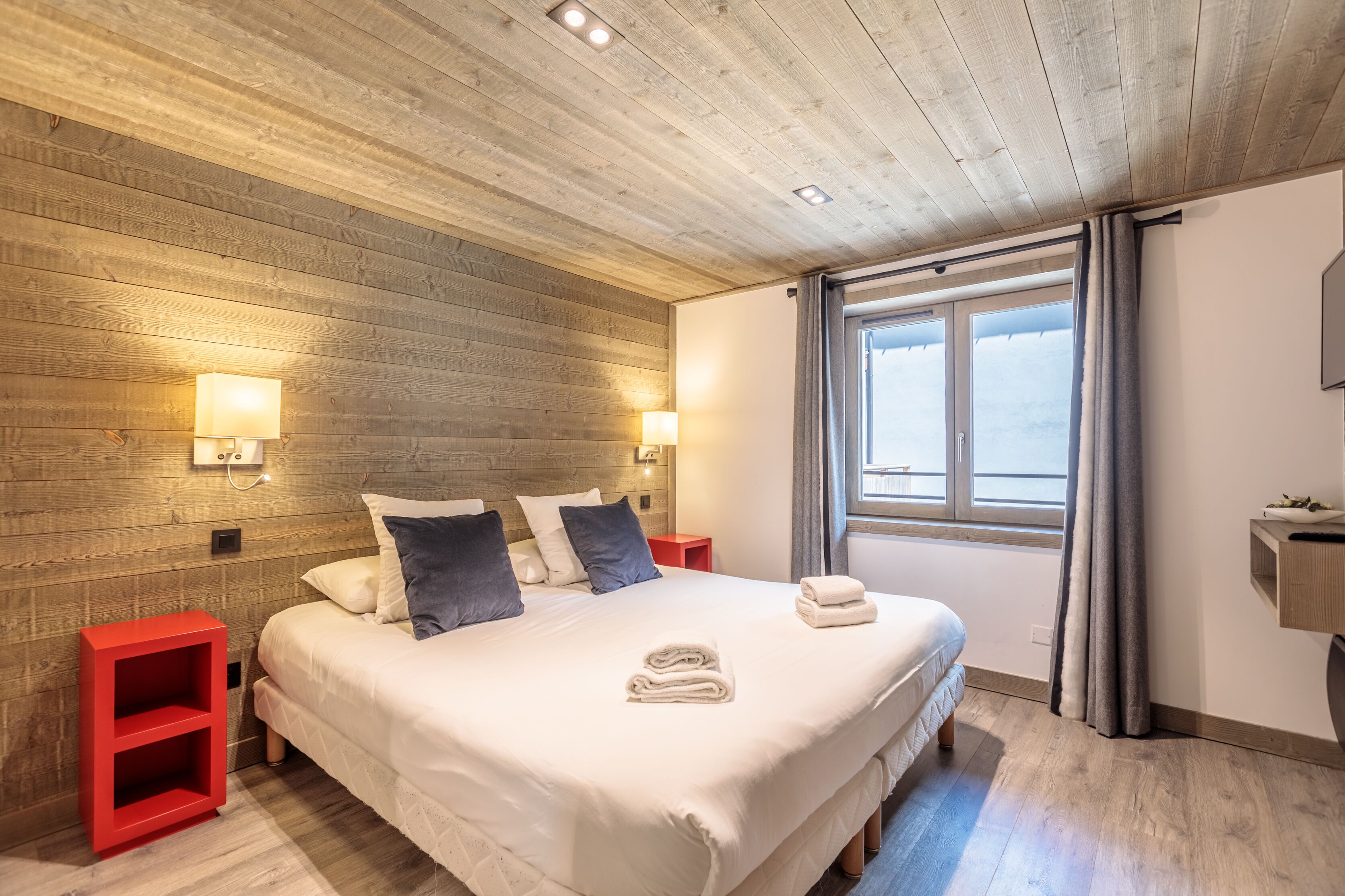 A bedroom with a wooden ceiling, a large bed with white bedding, two bedside lamps, and a window with curtains.