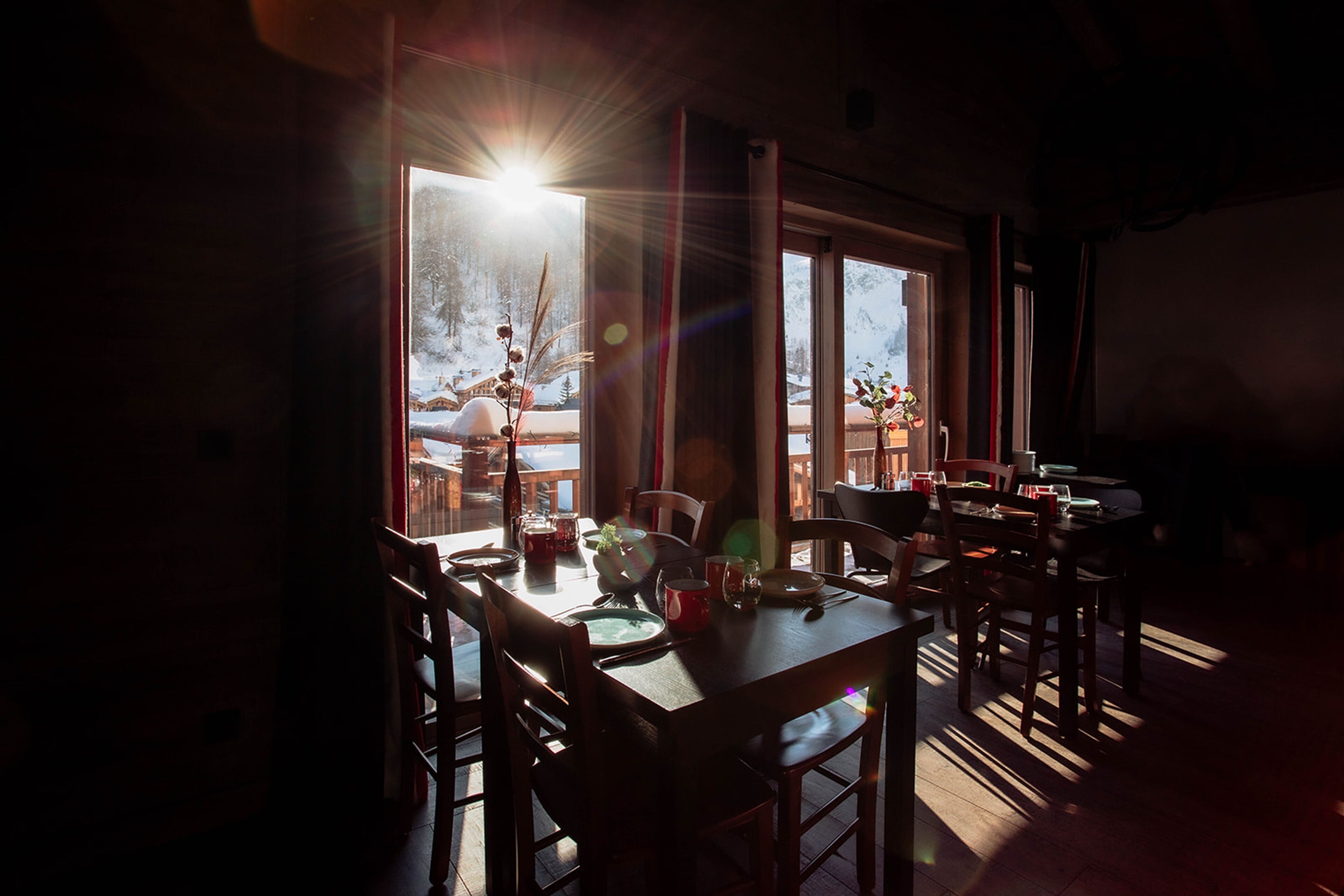 A dining area with a table set for two, large windows with a view of a snowy landscape, and a warm light source creating a cozy atmosphere.