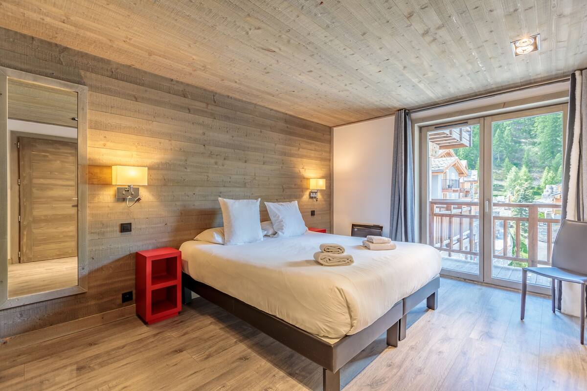 A modern bedroom with a wooden ceiling, a large bed with white linens, two bedside lamps, and a view of a wooden deck and trees through a large window.