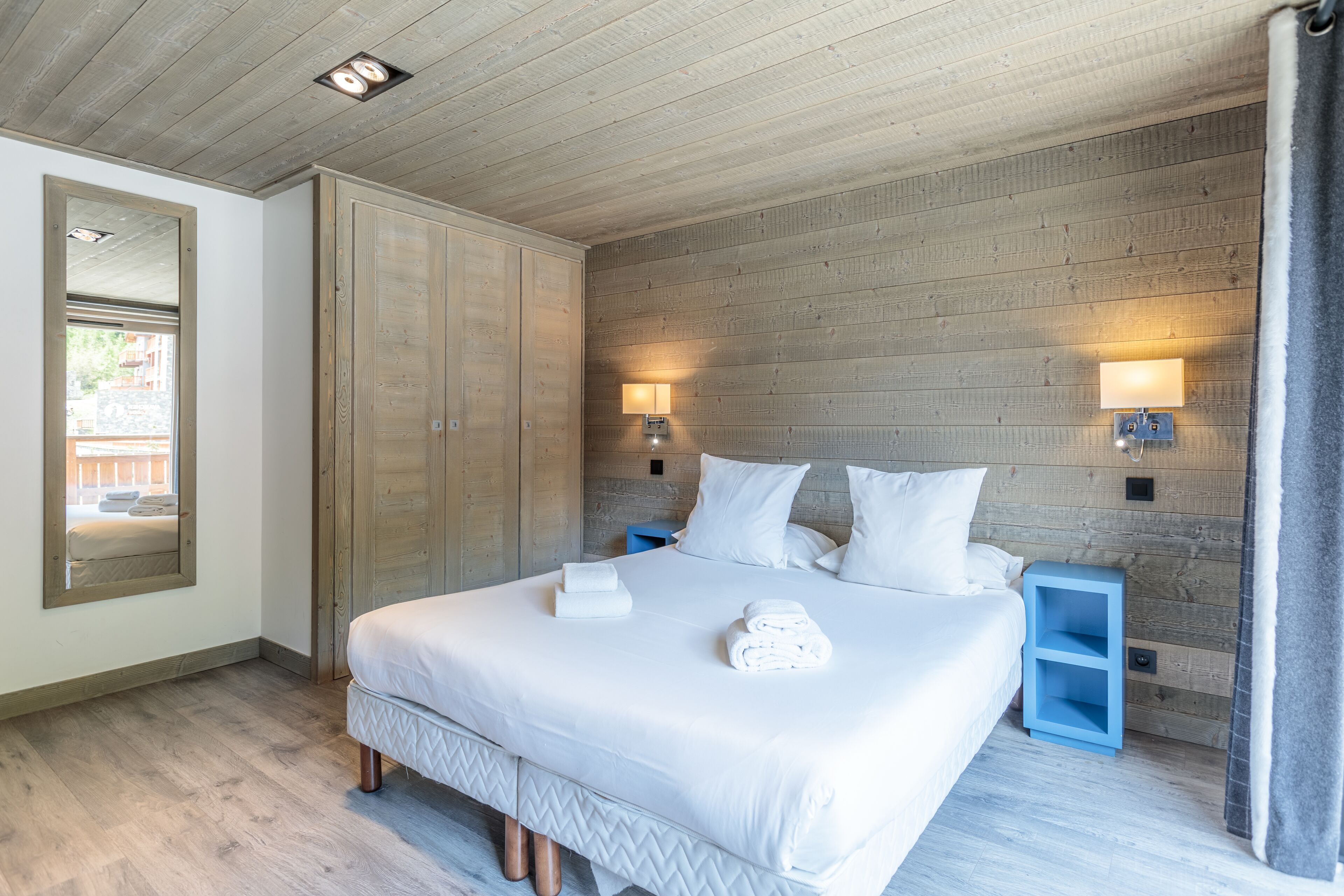 A bedroom with a large bed, wooden walls, two bedside lamps, a mirror, and a view of the outside.