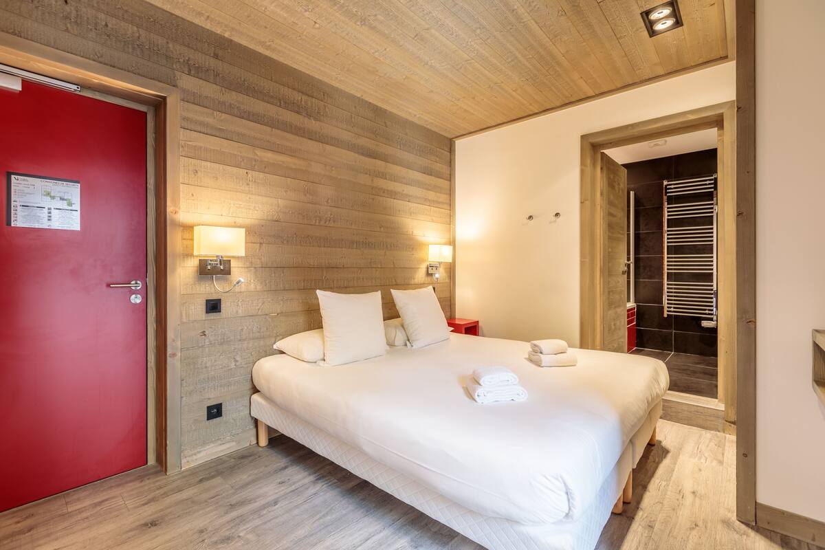 A hotel room with a wooden ceiling, a red door, a bed with white linens, two bedside lamps, and a towel on the bed.