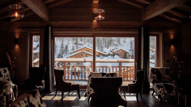 A cozy cabin interior with wooden walls, large windows, and a balcony overlooking a snowy landscape.