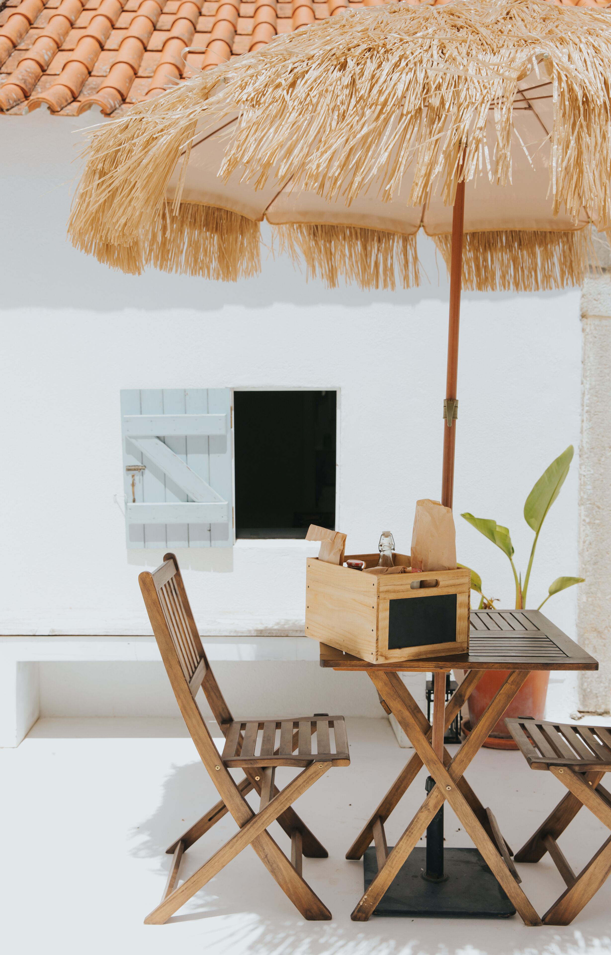 Una mesa y sillas de madera bajo un paraguas de paja, con una planta en maceta y una pequeña ventana en la pared.