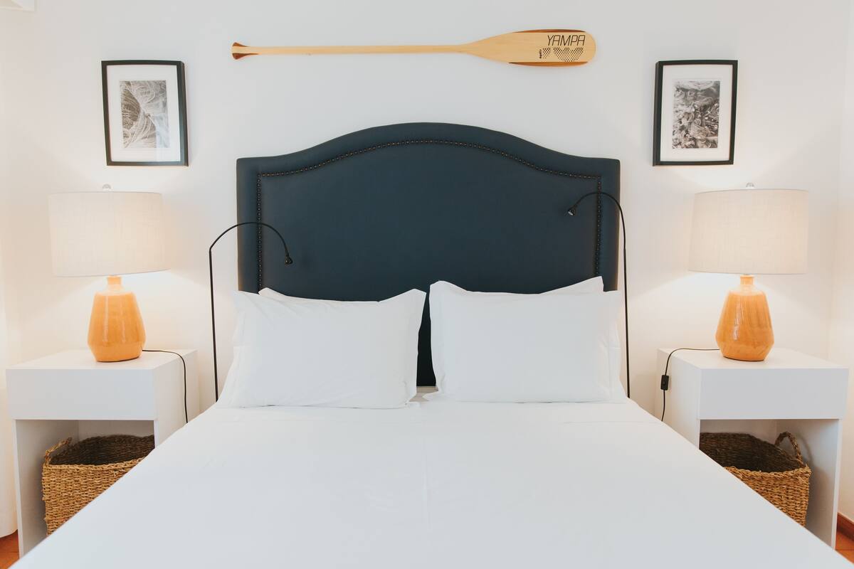 Una cama bien hecha con ropa blanca, flanqueada por dos mesitas de noche con lámparas y cestas.