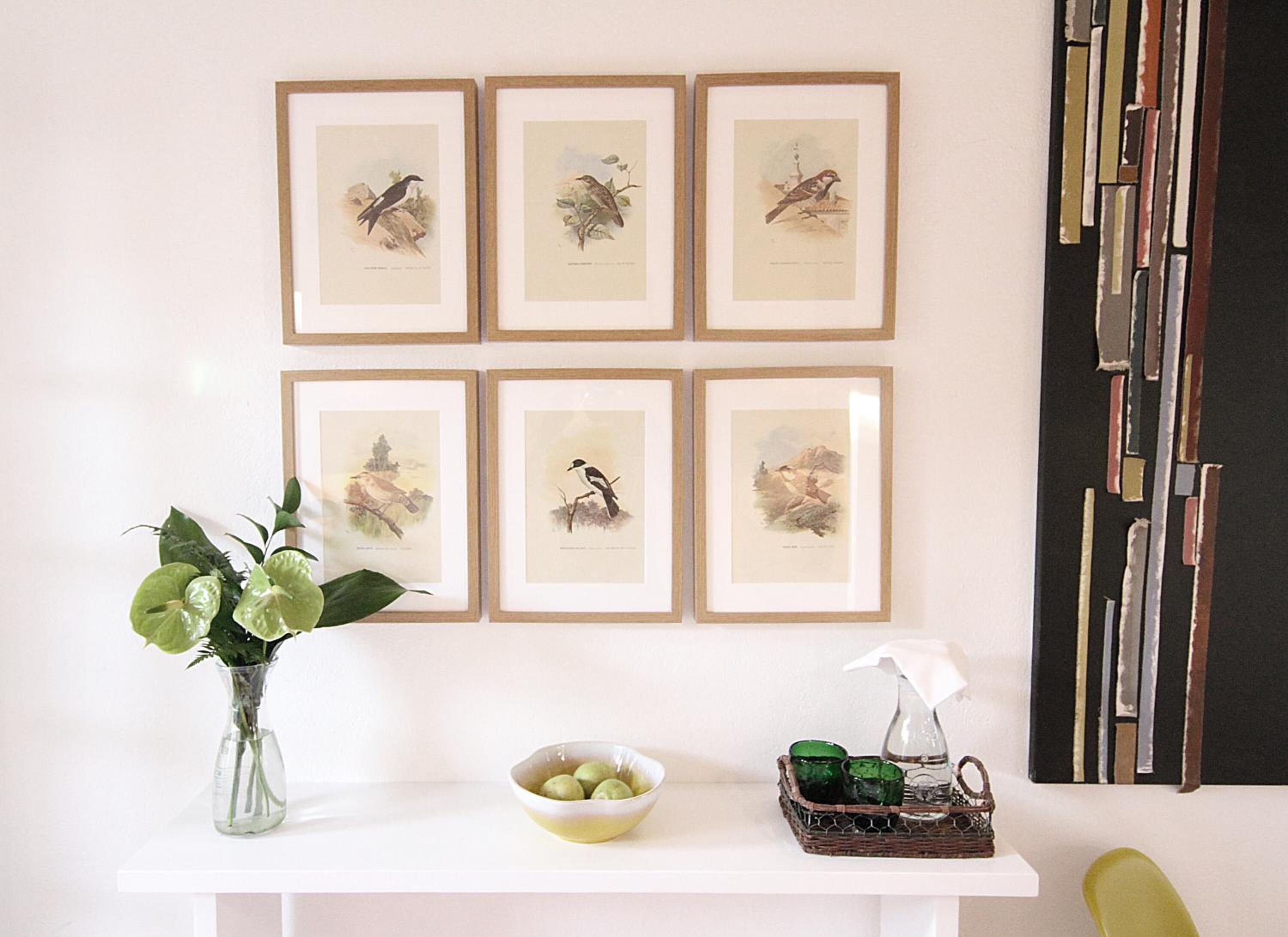 Une étagère blanche ornée de six illustrations d’oiseaux encadrées, un vase avec des feuilles vertes, un bol de pommes vertes et un panier en osier contenant des verres verts et un vaporisateur.