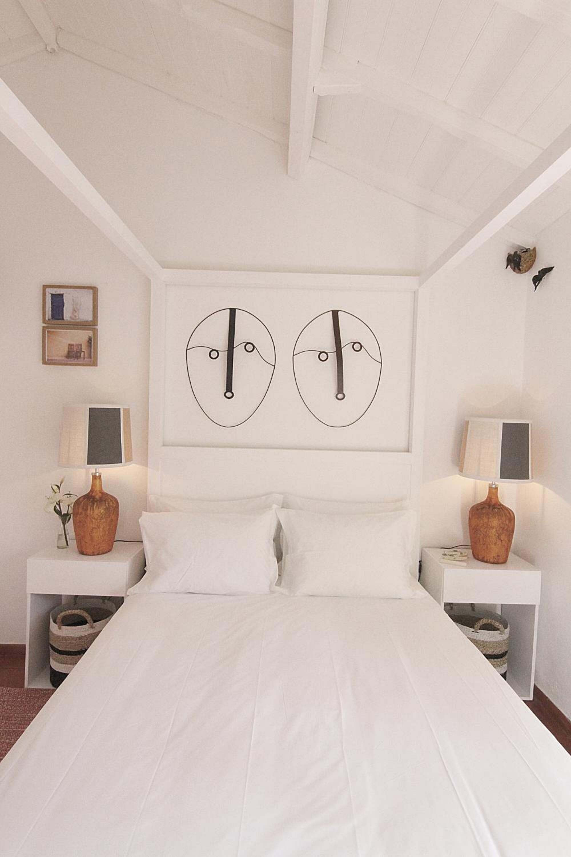 Un dormitorio con una cama blanca, dos mesitas de noche con lámparas y dos obras de arte abstracto en la pared.