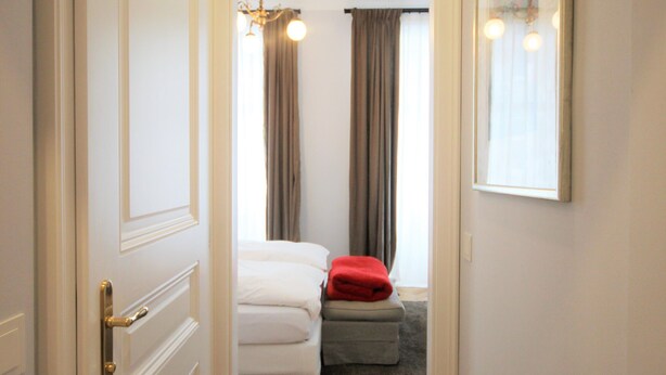 Ein Flur, der in ein Schlafzimmer mit einem Bett, einem roten Kissen und einem Fenster mit Vorhängen führt.