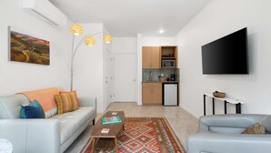 A modern living room with a sofa, coffee table, and TV. A kitchen is visible in the background with a microwave and cabinets.