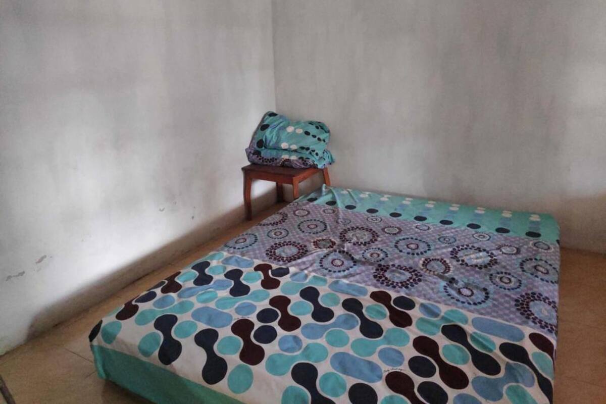 Una habitación pequeña y sencilla con una cama individual, un taburete de madera y una pared lisa.