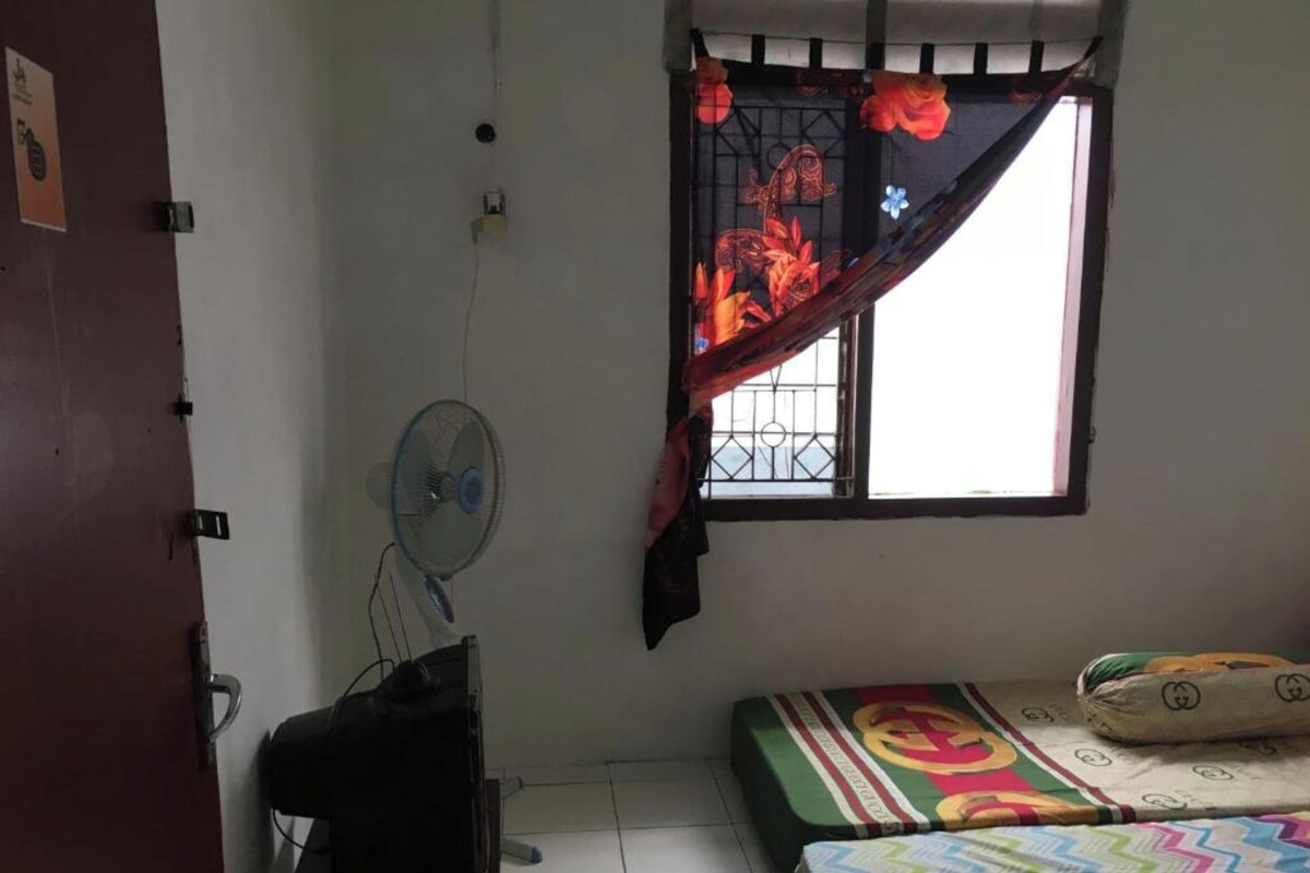 Una habitación pequeña y poco amoblada, con una cama, un escritorio, una silla, un ventilador y una ventana con cortina decorativa.