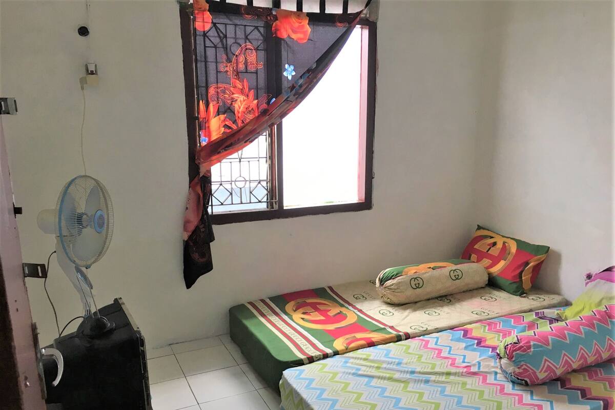A small, simple room with two beds, a window with a metal grill, a standing fan, and a wall socket.