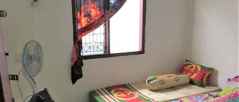 Una habitación pequeña y sencilla con dos camas, una ventana con reja de metal, un ventilador de pie y un enchufe en la pared.