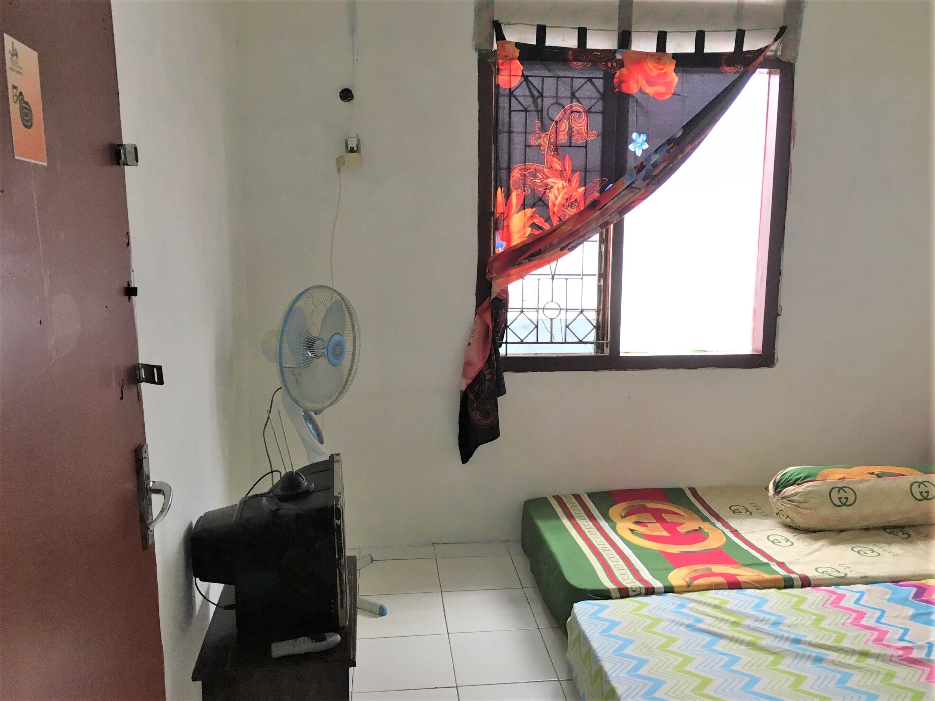 A small, sparsely furnished room with a single bed, a standing fan, a television, and a window with a decorative curtain.