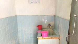 Une petite salle de bain carrelée, avec des toilettes à la turque, un lavabo en plastique rose et un seau rouge.