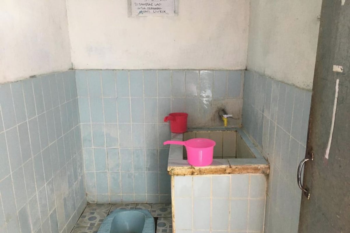 Un baño pequeño con un inodoro a piso, un cubo rojo y un grifo amarillo en la pared.