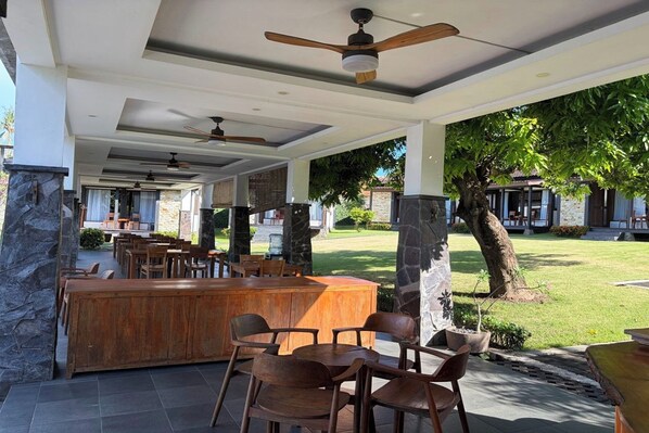 Un área de patio al aire libre con barra de madera, asientos, ventiladores de techo y vista a un césped y un edificio al fondo.