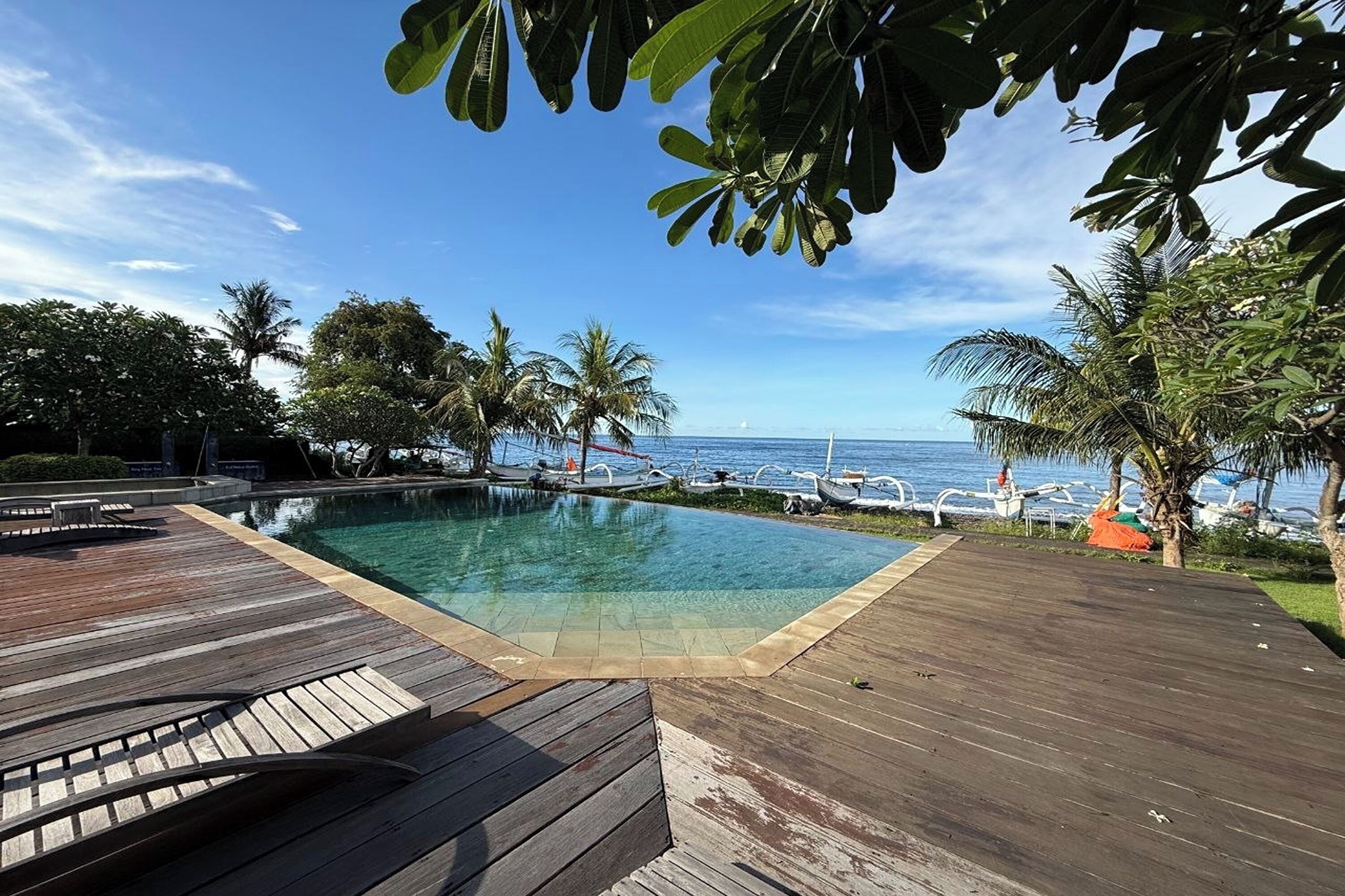 A wooden deck with a clear pool, palm trees, and a view of the ocean.