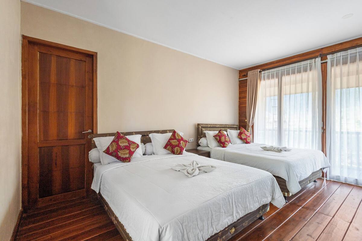 Een hotelkamer met twee bedden, houten vloeren, een grote houten deur en een raam met gordijnen.