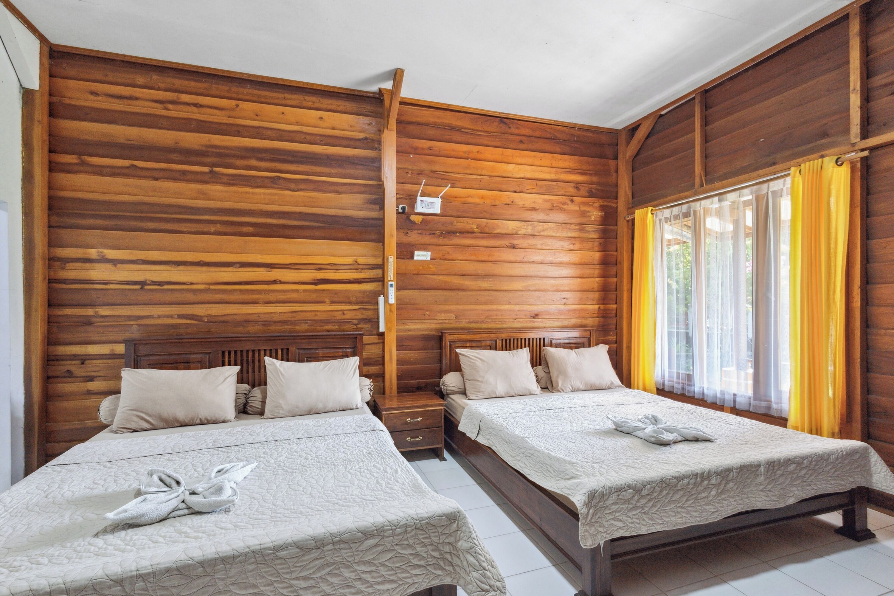 A room with two beds, wooden walls, a window with yellow curtains, and a small bedside table with a lamp.