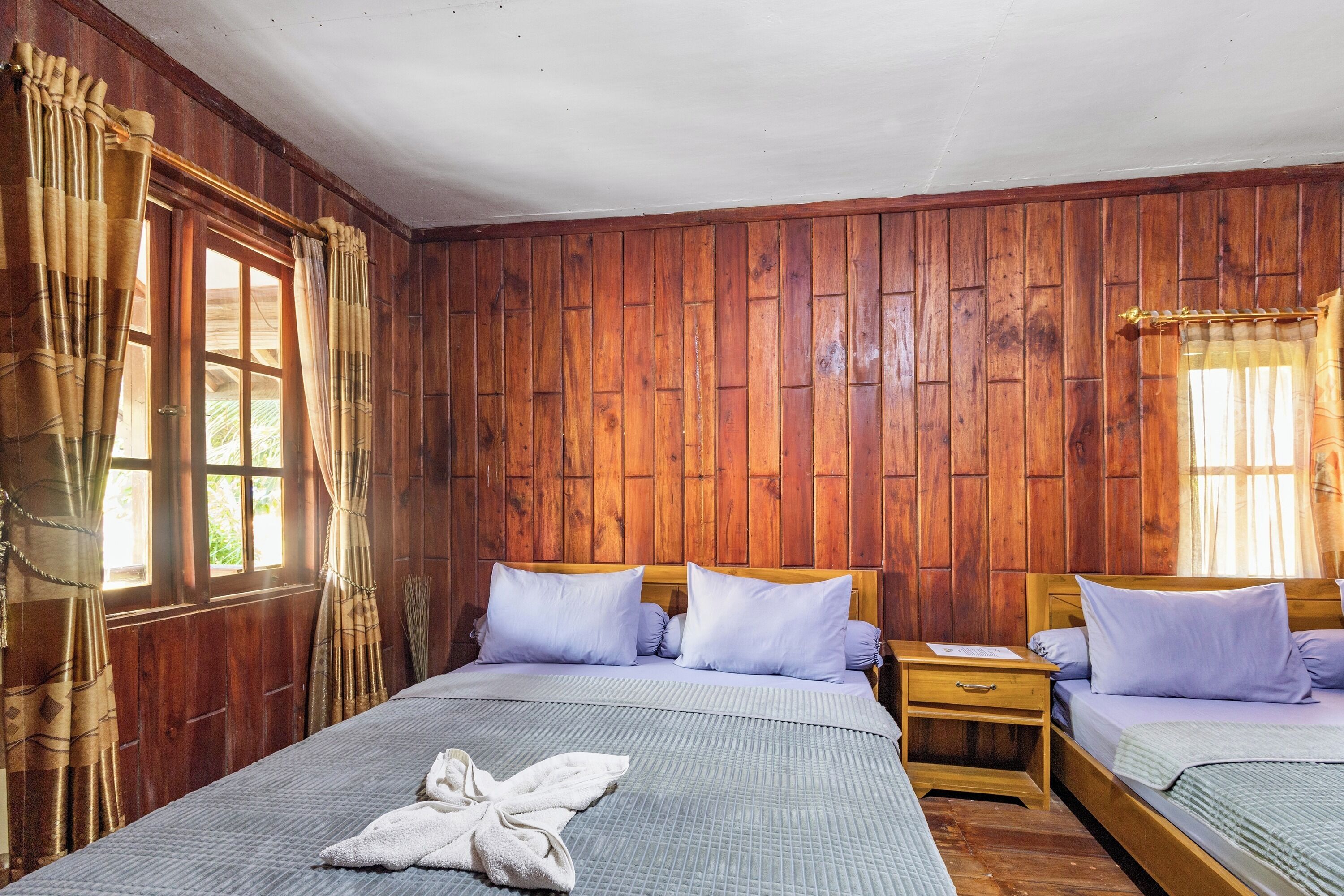 Quarto com painéis de madeira, duas camas, uma mesa de cabeceira e uma janela com cortinas.