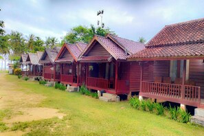 A row of wooden cabins with red roofs and balconies, situated on a grassy area with palm trees in the background.