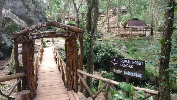 A wooden bridge with a sign indicating directions to 'Rumah Hobbit Puncak' and 'Kebersihan'. - Seribu Batu Songgo Langit Resort & Camp (Mangunan)