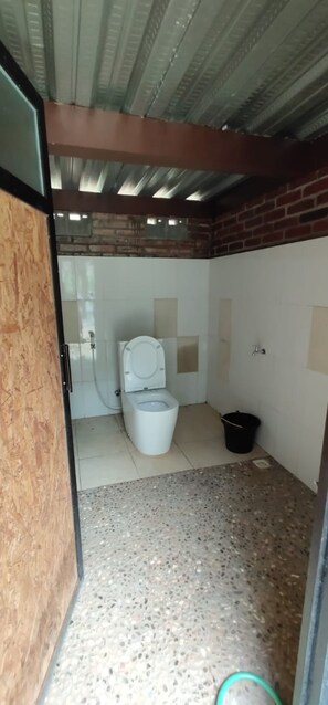 Une petite salle de bain fermée, comprenant des toilettes, un seau noir et un sol carrelé.
