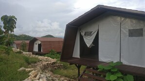 A row of rustic wooden cabins with white curtains, set in a lush green landscape with a stone path leading to them. - Watu Mabur Holiday Centre and Camping (Mangunan)