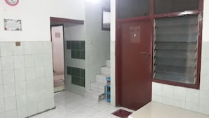 A narrow hallway with a tiled floor, a red door with a window, and a small blue container on the floor. - Homestay Mas Arjuna - Female Only (Surabaya)