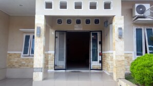 A building entrance with a tiled floor, a set of steps leading up to the door, and a small green bush to the right. - Dedy Kost (Jambi)