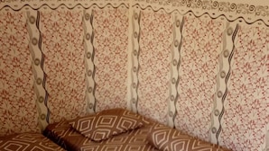 A bed with a geometric patterned bedspread and pillows, a floral wallpaper headboard, and a tiled floor. - Dedy Kost (Jambi)