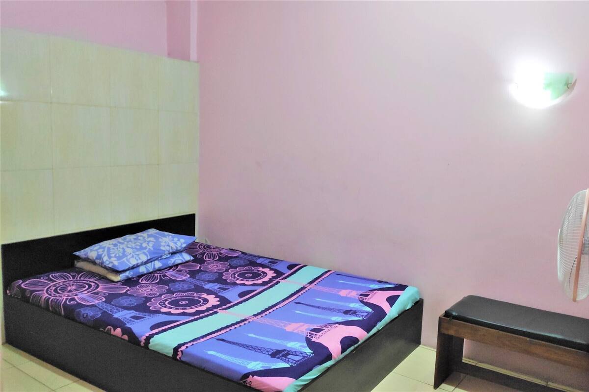 A single bed with a patterned bedspread, a bedside table, a wall-mounted fan, and a light fixture.