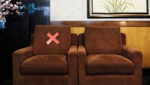 Un canapé marron orné d’un coussin sur lequel est dessiné un « X » rouge, placé contre un mur où sont accrochés un tableau encadré et un vase de fleurs.