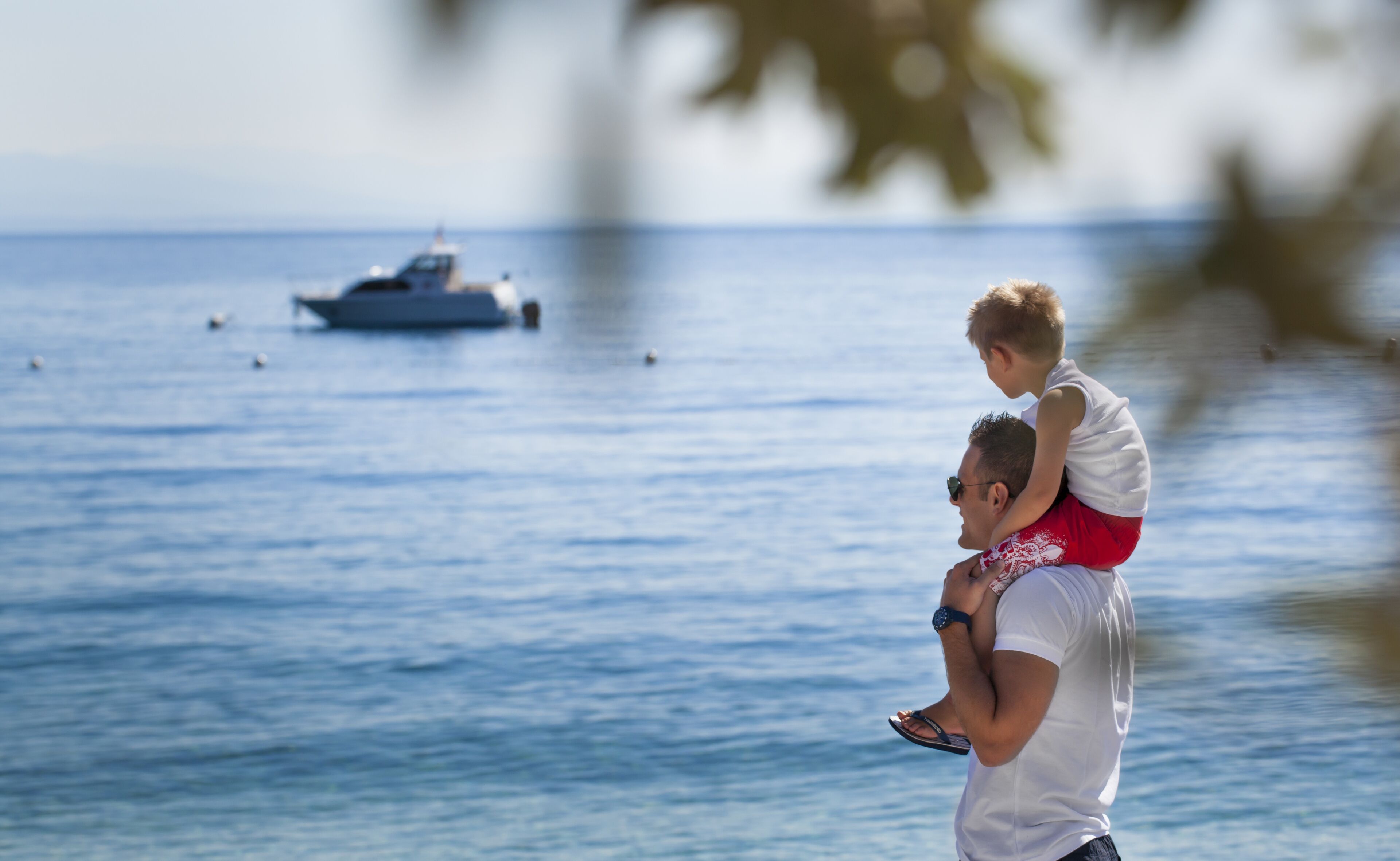 Un hombre lleva a un niño sobre sus hombros junto a un cuerpo de agua, con un barco a lo lejos.
