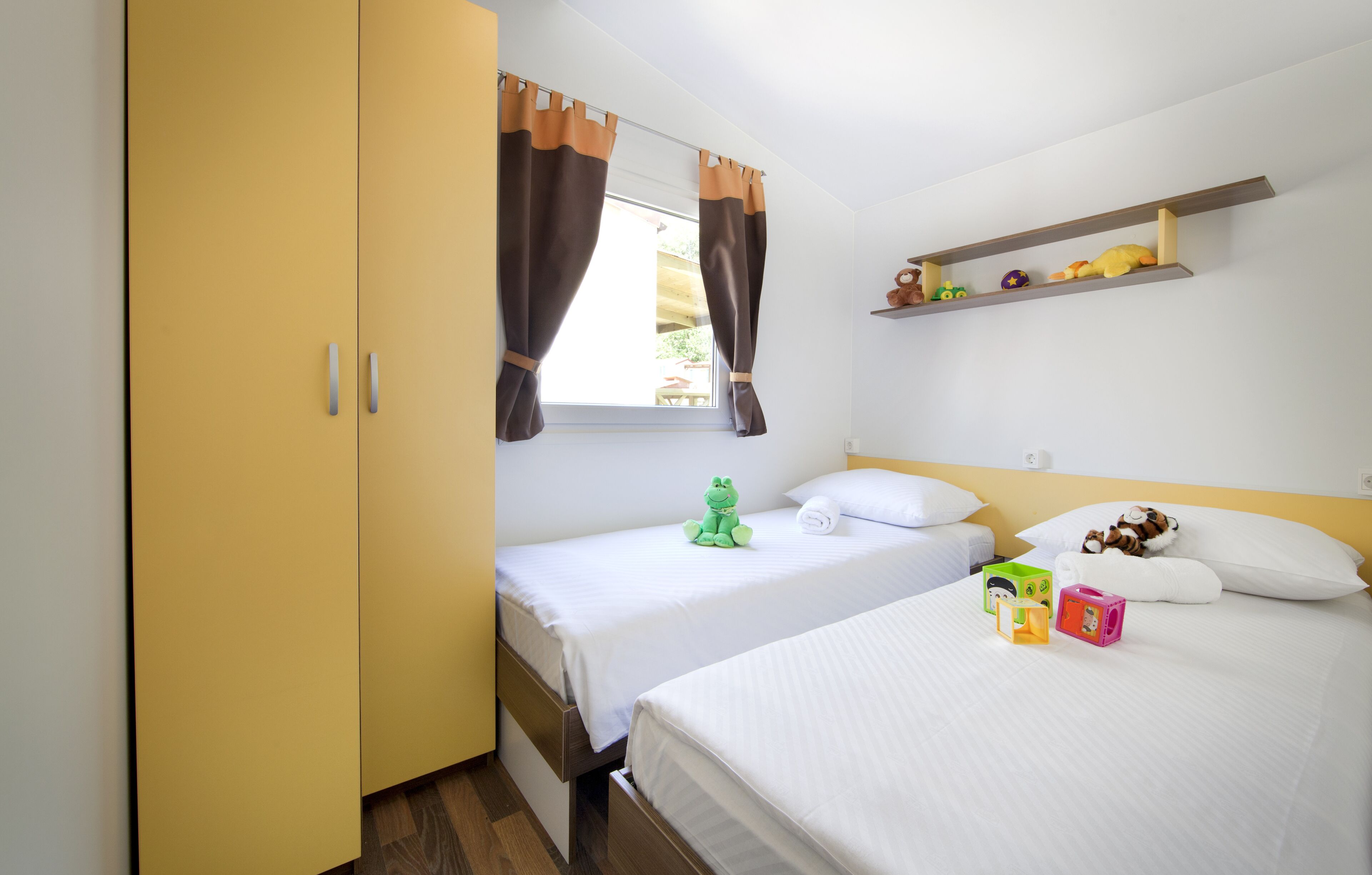 Un dormitorio con dos camas, un armario, una ventana con cortinas y una estantería con juguetes, además de un sapo de juguete verde sobre una de las camas.