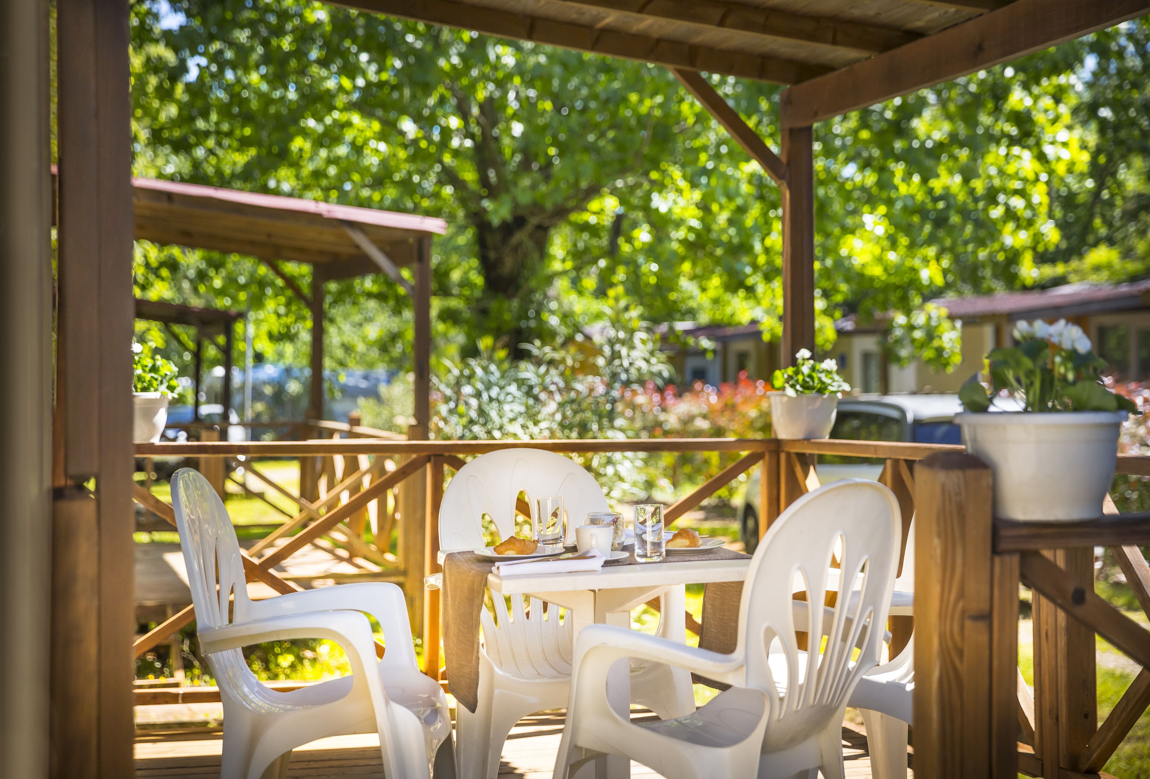 Une terrasse extérieure avec des tables en bois et des chaises en plastique blanc, entourée de plantes en pot et de verdure.