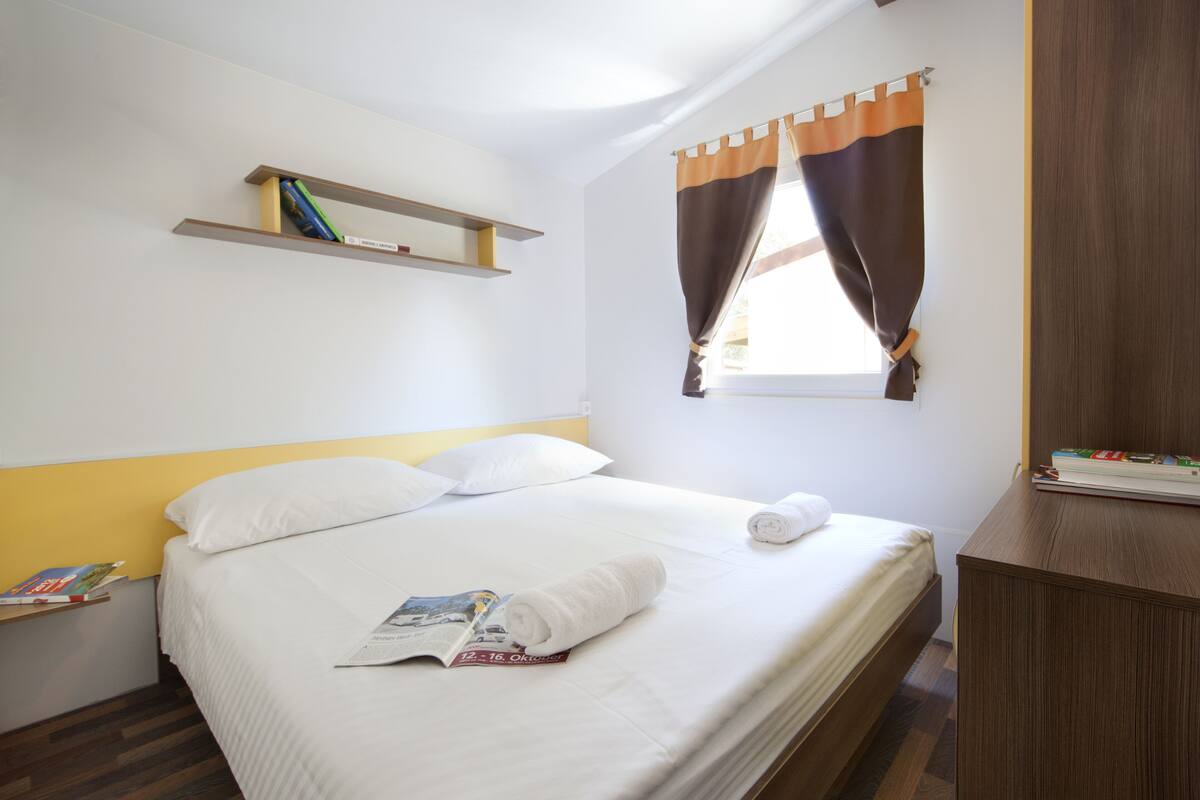 A neatly made bed with pillows and a rolled towel, a small shelf with books, and a window with curtains.