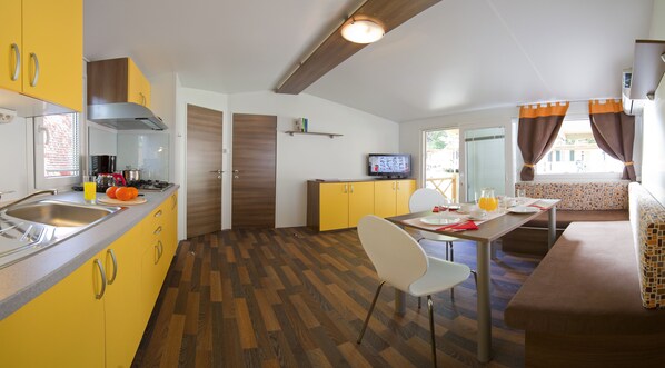 Cocina compacta con un lavamanos, estufa y armarios amarillos. Hay un comedor con mesa y sillas, y un televisor al fondo.