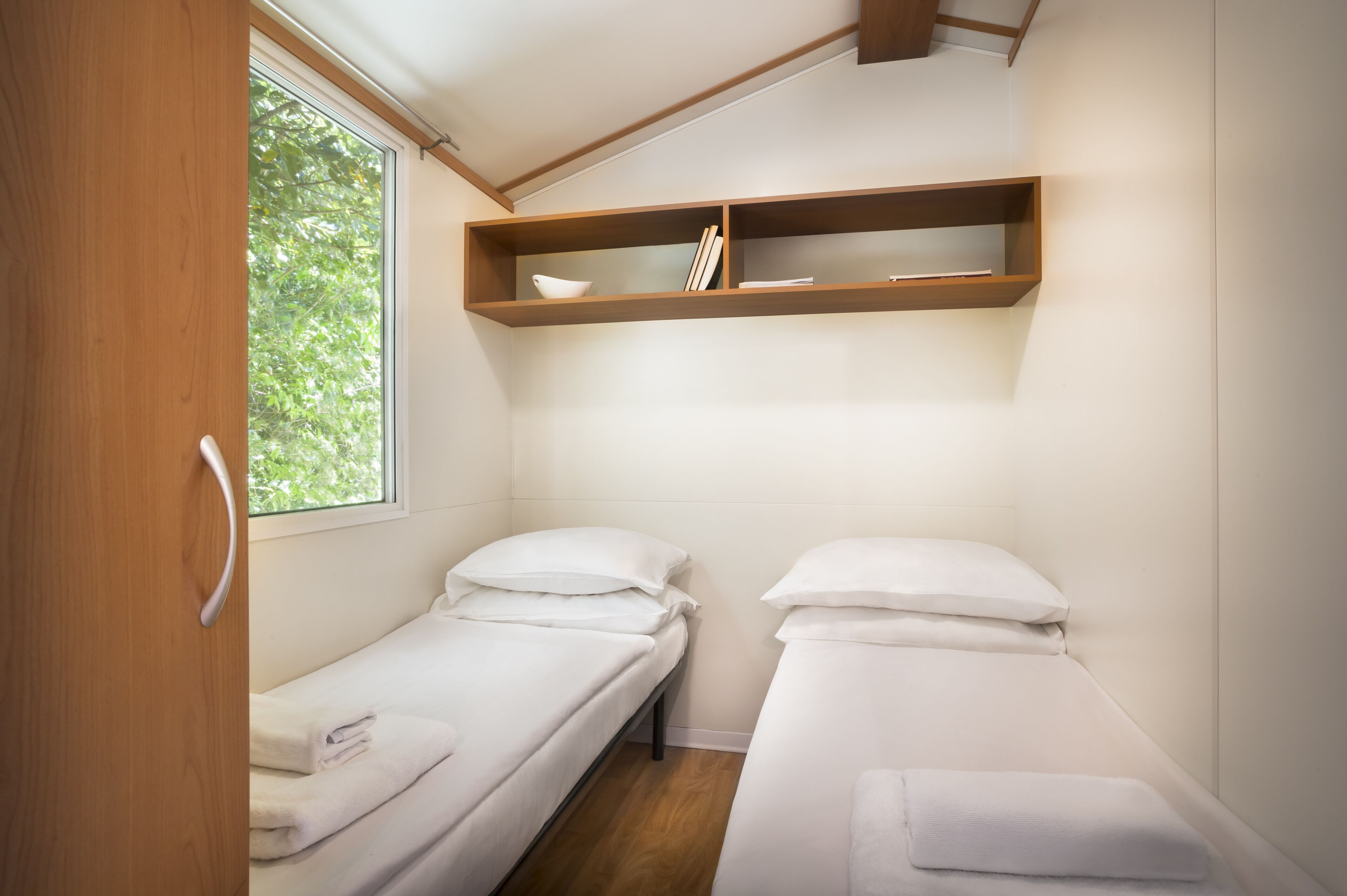 A small, compact room with two single beds, a wooden shelf above, and a window providing natural light.