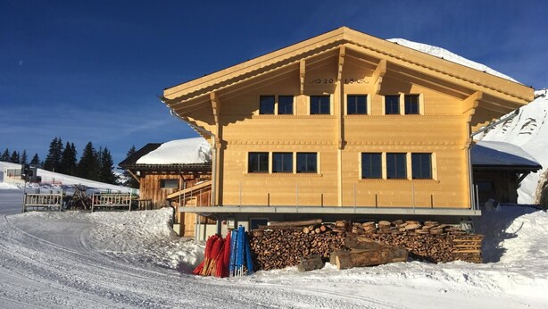 Un chalet de madera de dos pisos con techo inclinado, ventanas grandes y un paisaje de montaña nevada al fondo.