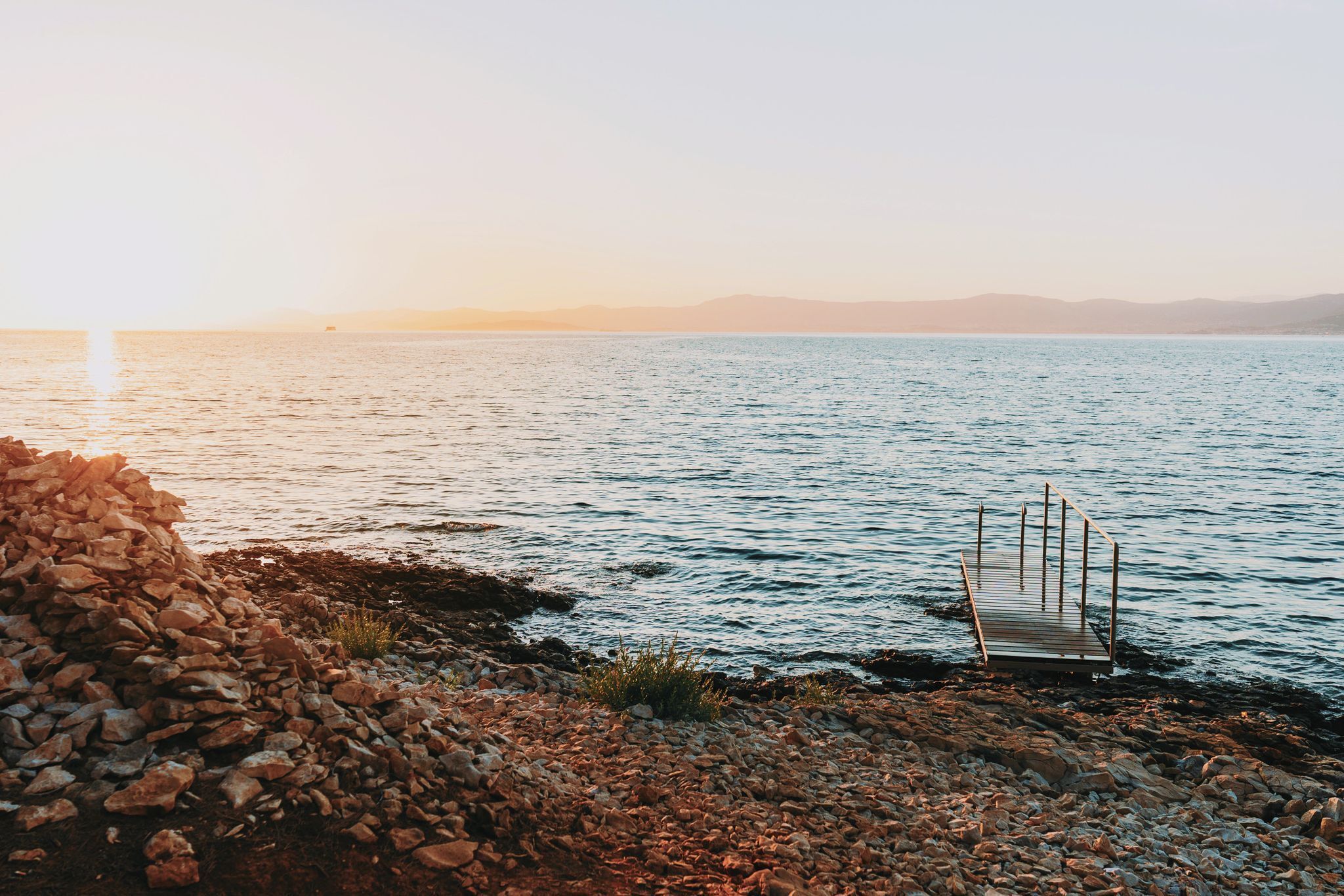 A rocky shoreline with a wooden dock extending into calm waters, surrounded by a clear sky.