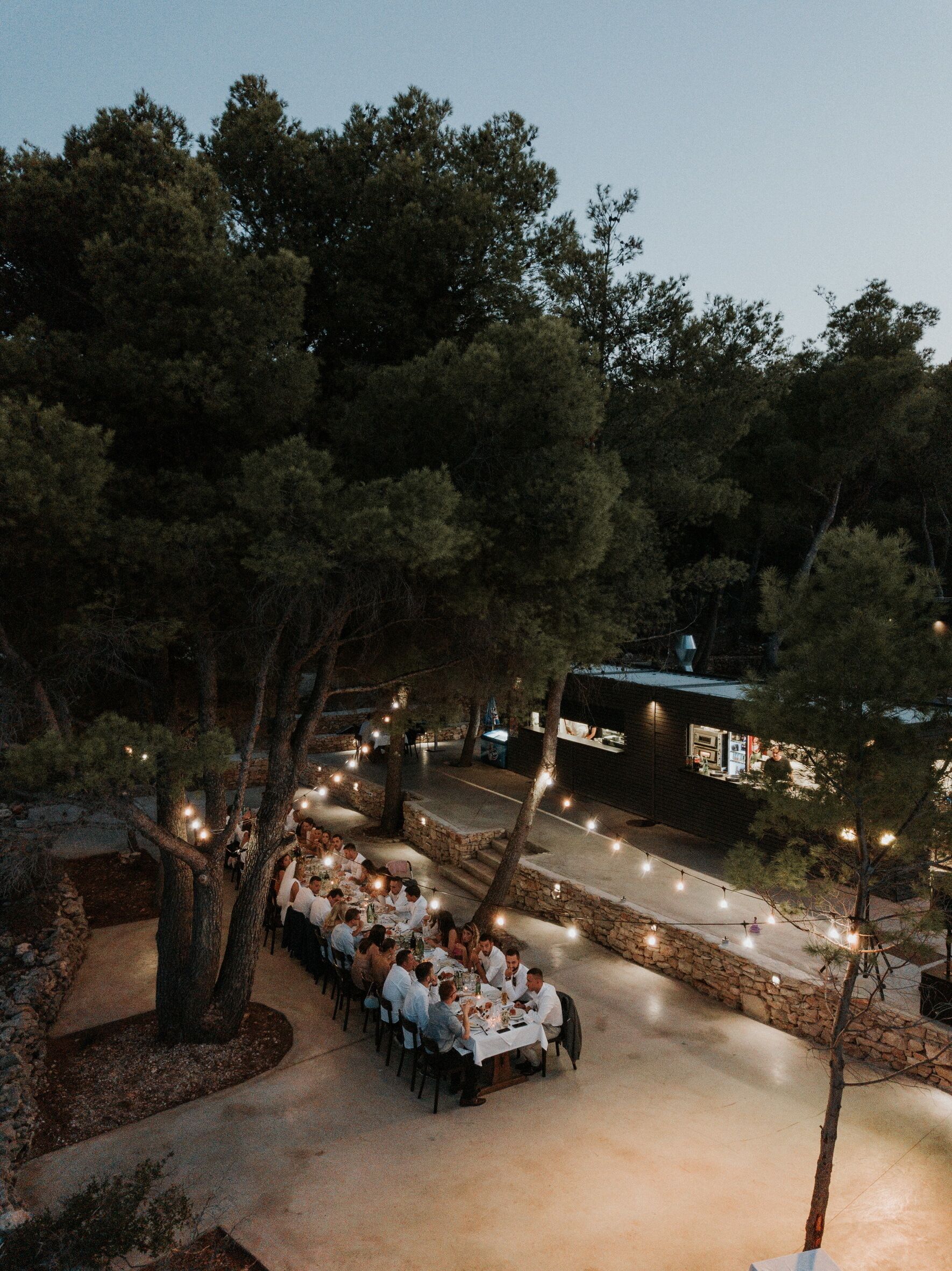 Un área de comedor al aire libre con mesas dispuestas bajo árboles, rodeada de muros de piedra y luces colgantes.