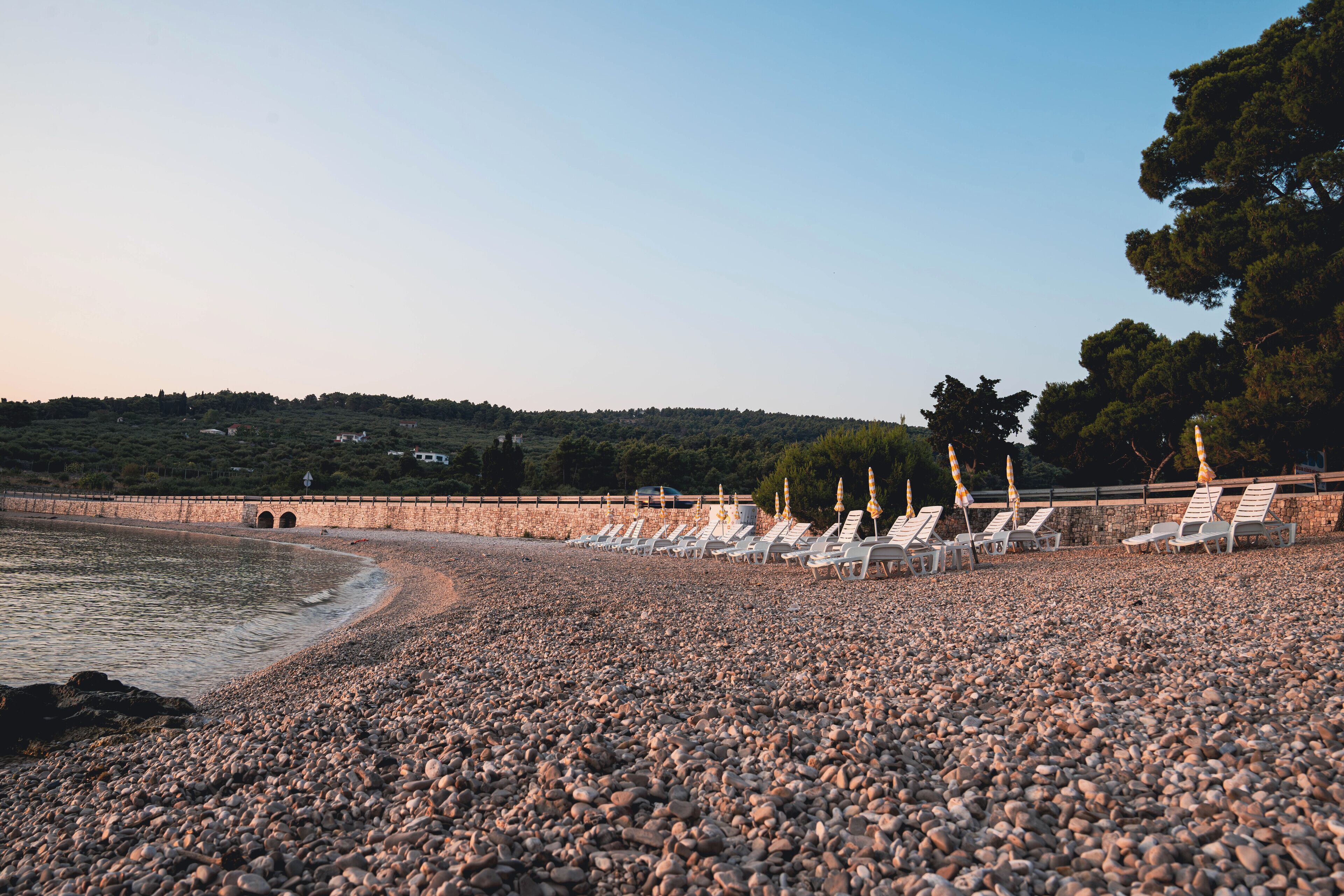 A pebble beach with lounge chairs and umbrellas, a stone wall, and a forested hill in the background.