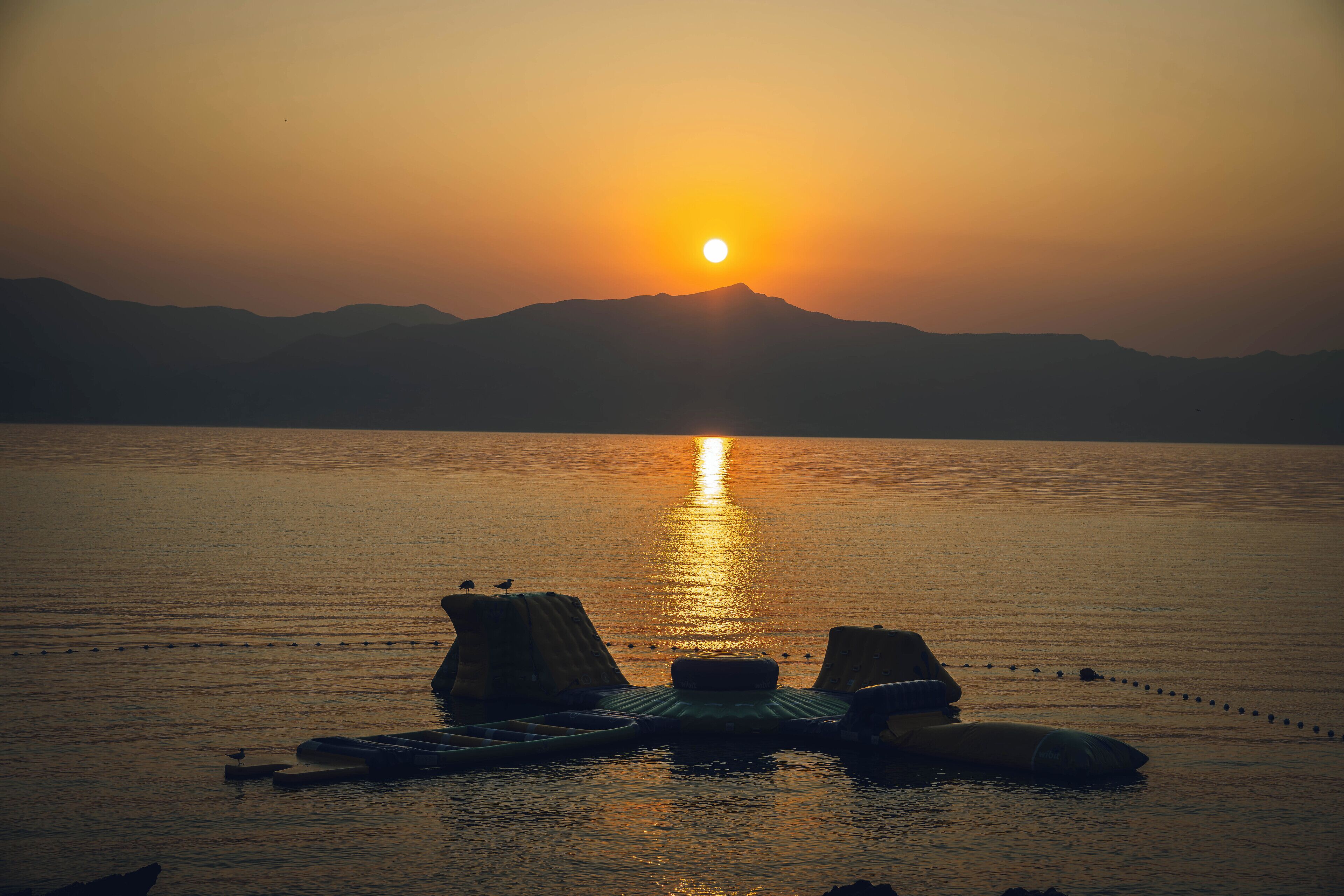 A sunset over a calm body of water with mountains in the background and floating platforms with seating.