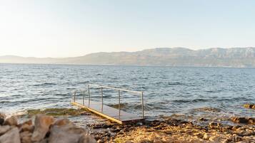 A rocky shoreline with a wooden dock extending into calm water, surrounded by mountains in the background.