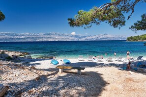 A pebble beach with lounge chairs and a wooden bench, overlooking a clear blue sea with mountains in the background. - Boutique Camping Bunja (Supetar)