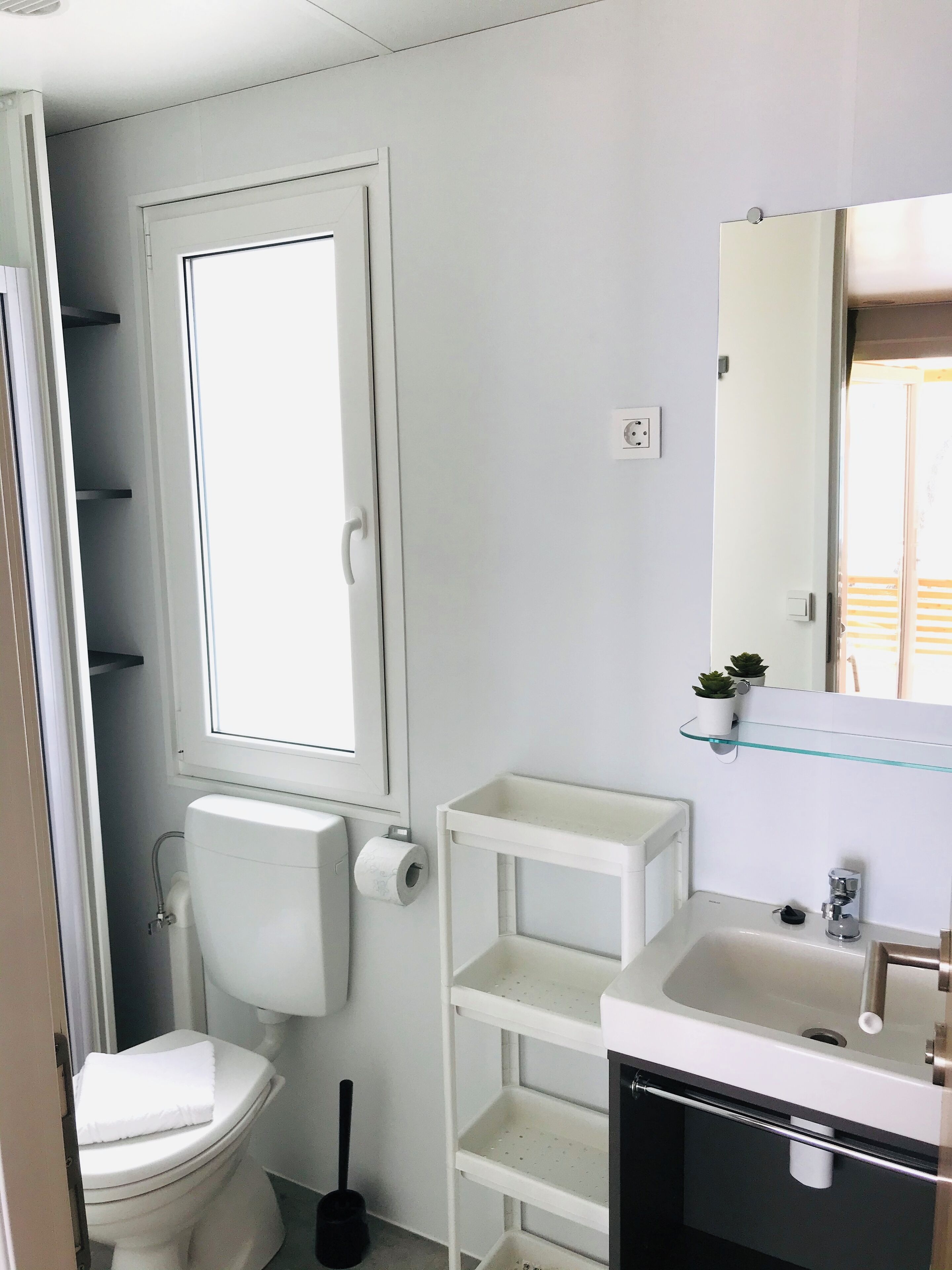 A small, compact bathroom with a toilet, a glass shelf above the sink, and a window.