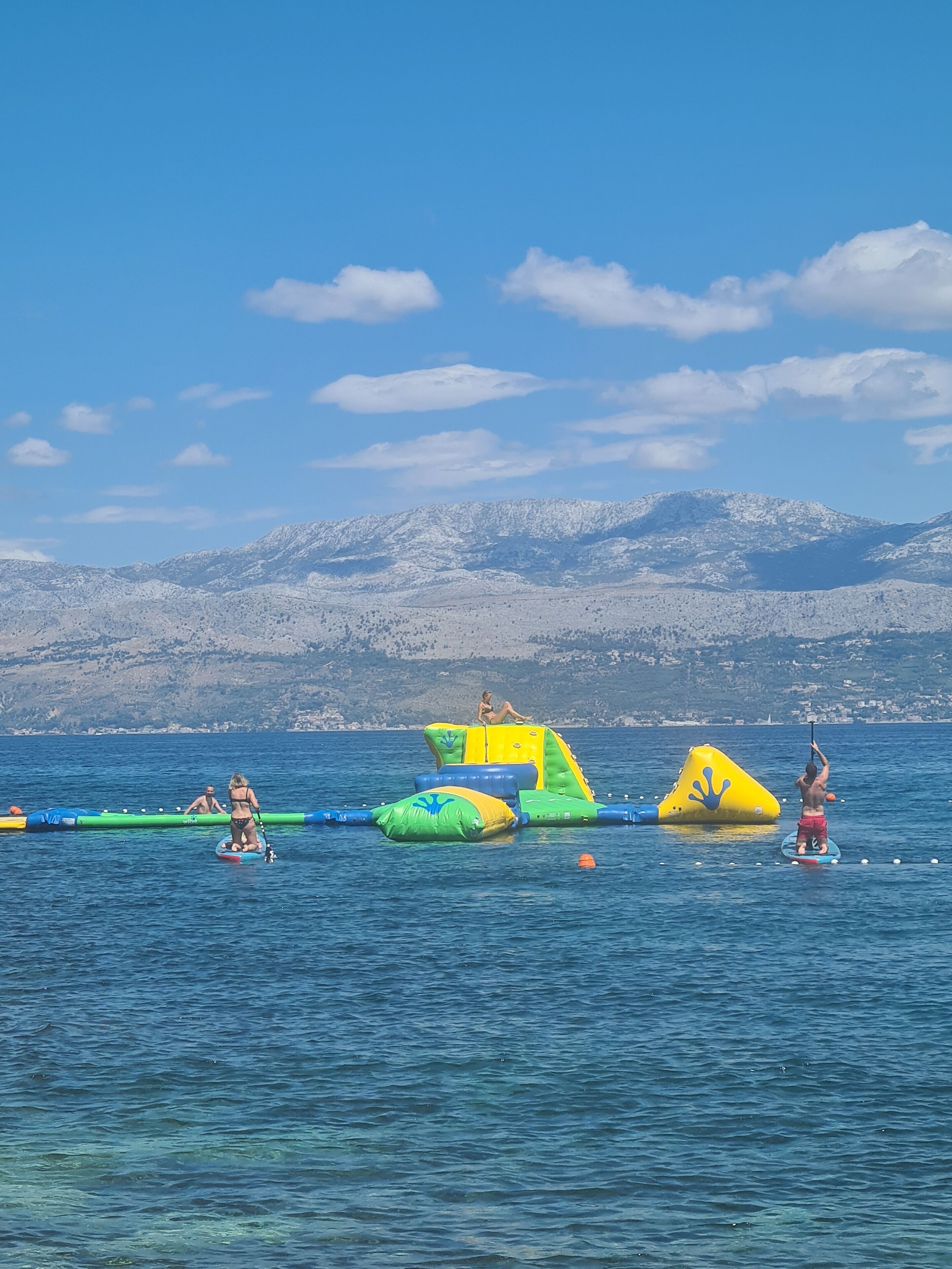 Escena de playa con juguetes inflables para agua y gente en tablas de paddle.
