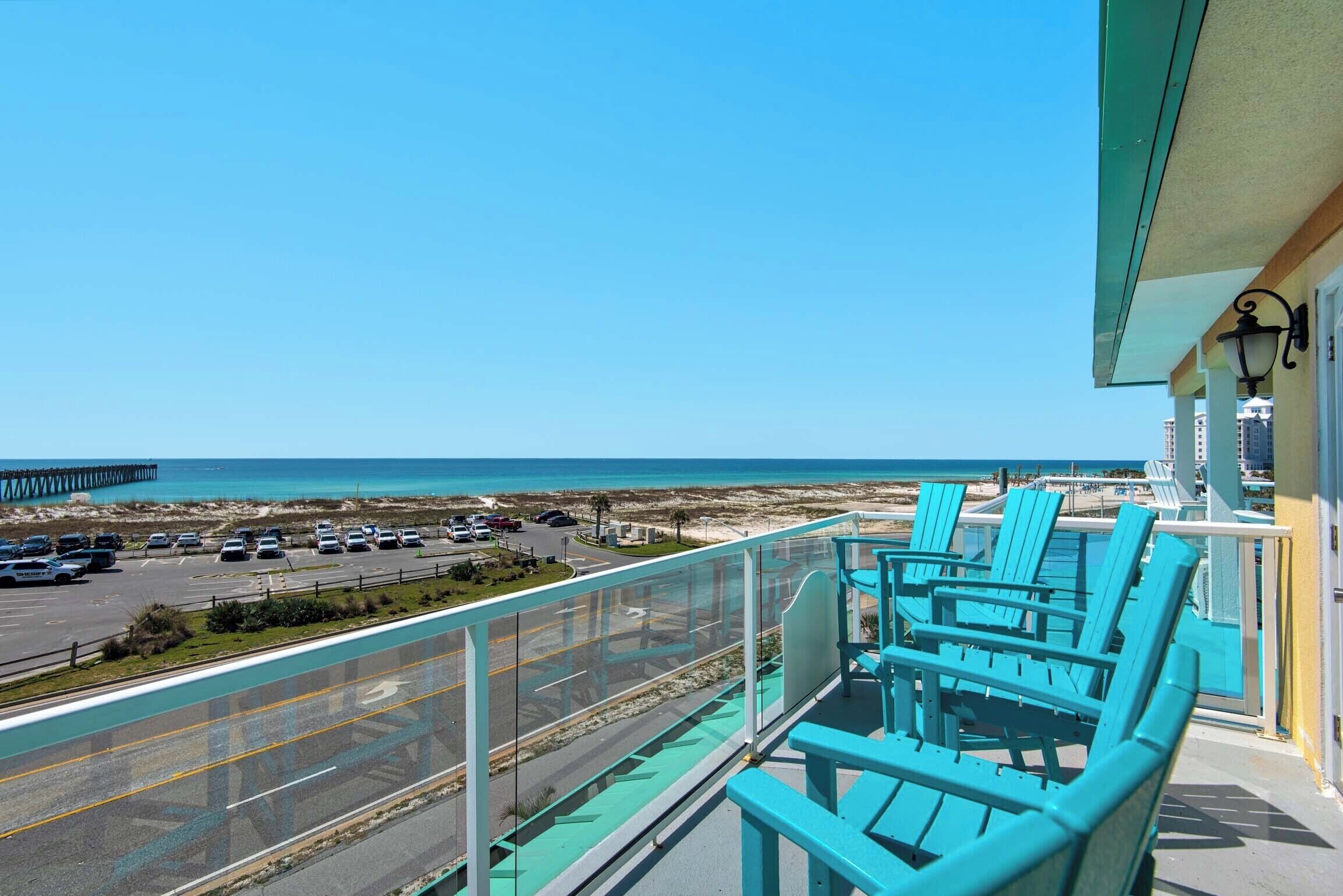 A balcony with turquoise chairs overlooking a beach and a parking area.